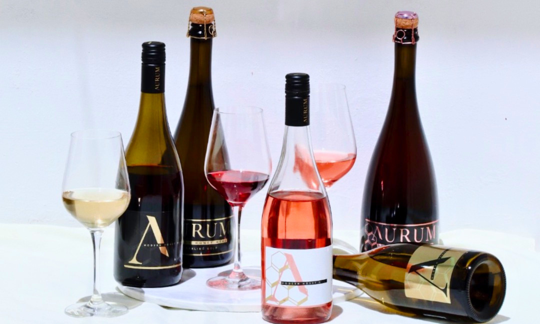 The AURUM range. Four bottles of award-winning Australian honey wine with poured glasses of white, red and rosé, styled on a white marble surface.
