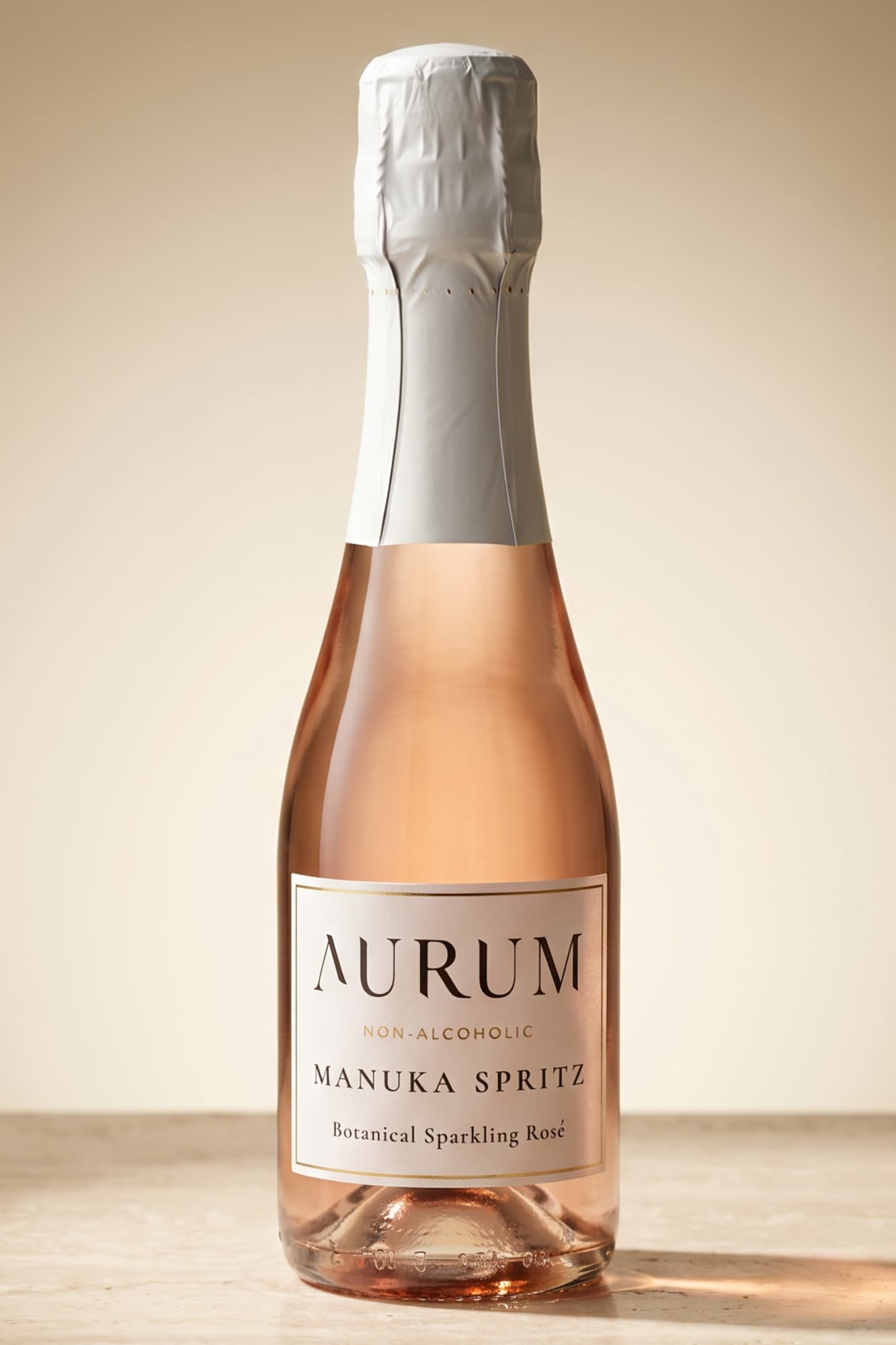 Aurum Manuka Spritz non-alcoholic sparkling rose wine in a clear glass mini champagne bottle with a white foil capsule and a cream-colored label, set against a warm beige background.